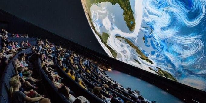 Imagem mostra pessoas sentadas, em sala parecida com a de cinema, e universo sendo projetado em planetário, em Miami. 