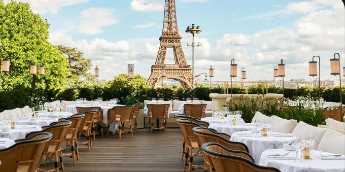 Imagem de mesas de restaurantes em área externa, com a Torre Eiffel ao fundo, em Paris