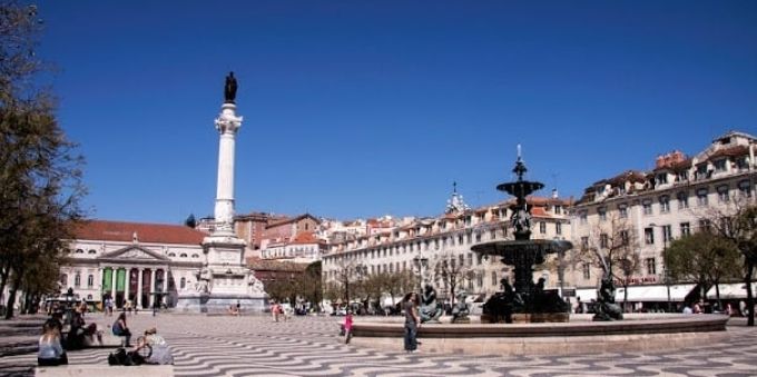 Imagem mostra praça ao céu aberto, com monumento e fonte de água, em Lisboa 