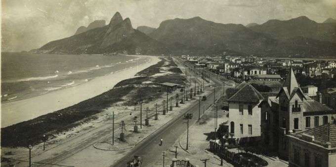 Na imagem temos a fotografia da região da praia de Ipanema no passado. A foto é em preto e branco e é possível ver a a praia, o pão de açúcar, e algumas casas.