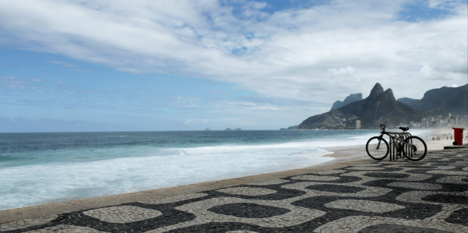 Na foto é possível ver o calçadão da praia de Ipanema com uma bicicleta ao fundo. É possível ver um pouco do mar a esquerda e bem mais distante montanhas e uma paisagem verde.